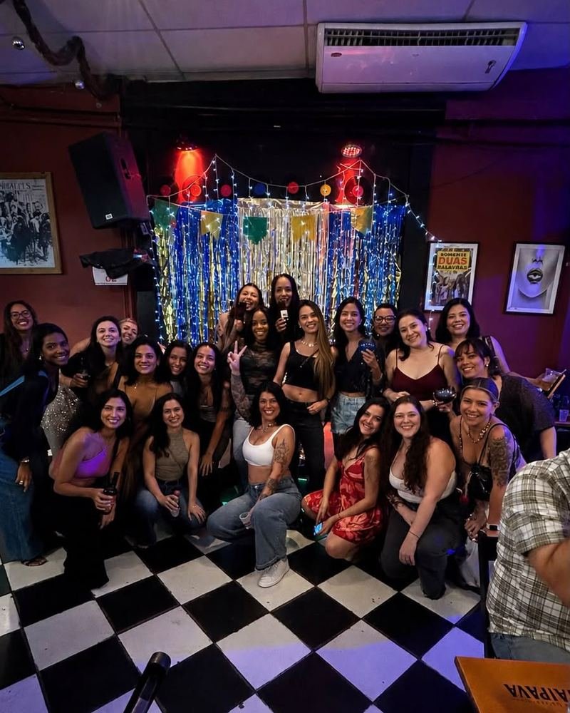 Grande grupo de participantes reunidas para a foto em uma noite de karaokê, diante de uma cortina metálica com luzinhas e bandeirinhas.