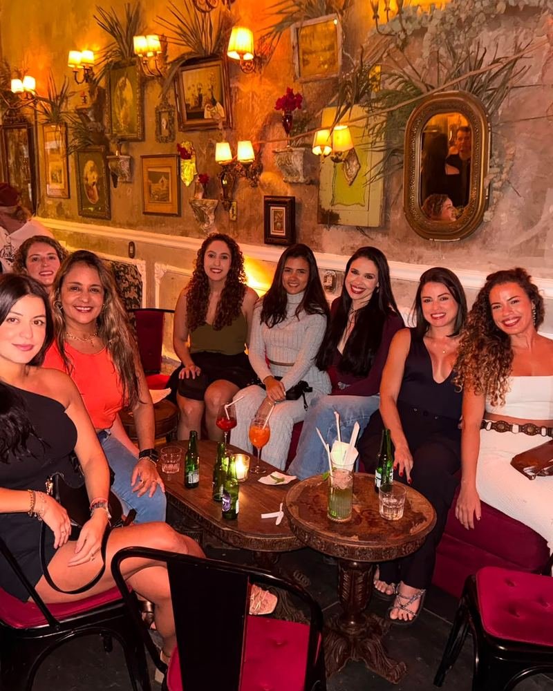 Nove participantes sentadas em volta de mesinhas de madeira em um restaurante de luz baixa, com quadros e espelhos antigos cobrindo a parede.