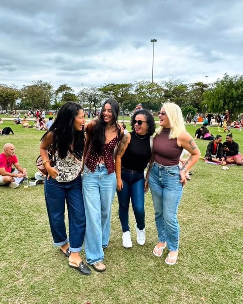 Quatro participantes rindo juntas, de braços dados, no gramado durante a Feira da Glória, com outras pessoas sentadas na grama ao fundo.